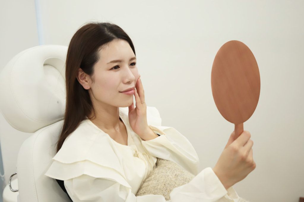 頬に手を当てながら、手鏡で自身の顔を見てほほえむ女性